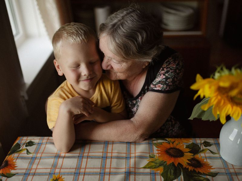An older woman hugs her young grandson at a kitchen table beside a vase of sunflowers.