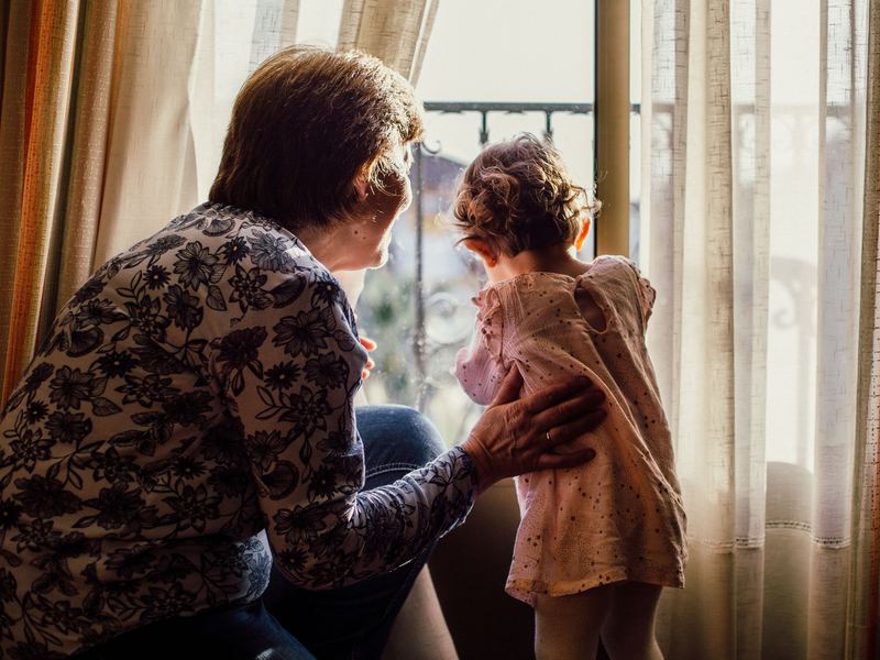 A grandmother kneels beside a toddler as they look out of a sunlit window together.