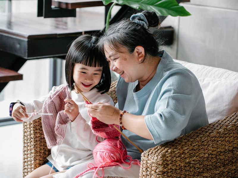 A smiling grandmother helps her granddaughter learn to knit in a bright living room.