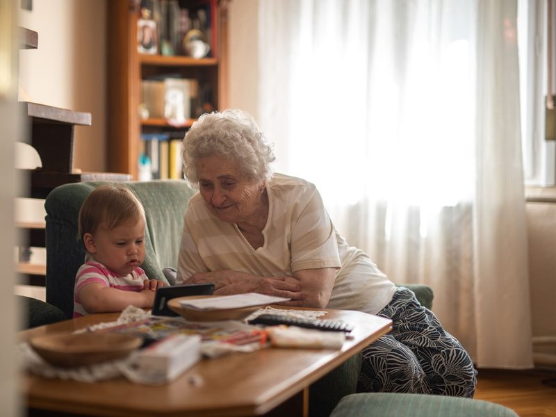 An elderly woman and a baby sit together at a low table covered with papers and drawings.